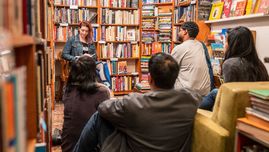Na zdjęciu troje ludzi przysłuchujących się wypowiedzi kobiety trzymającej książkę i siedzącej przed nimi. W tle znajdują się regały z książkami.